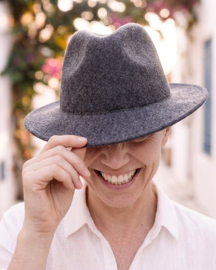 A smiling person in a white linen shirt tips a grey felt hat down over their eyes on a sunlit street with bougainvillea.